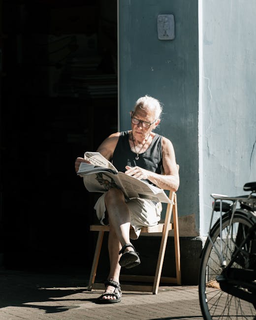 A senior woman with short gray hair and glasses is seated on a wooden chair outdoors beside a light blue building, reading a newspaper. She is dressed in a black sleeveless top and beige shorts, with
